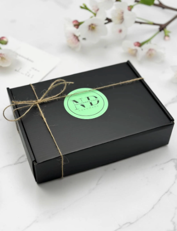 Black box with a green label tied with string on a white surface with flowers in the background