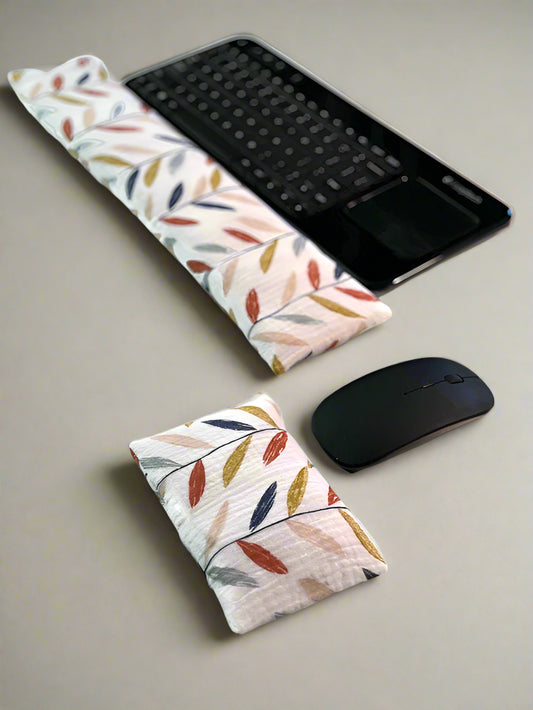 A black wireless keyboard with a built-in touchpad is placed on a white surface. In front of it lies a long, colorful leaves cream fabric wrist rest. Nearby, a white wireless mouse is accompanied by a smaller, matching wrist rest for ergonomic support.