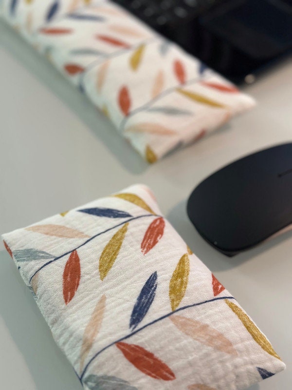 Cream color fabric wrist rest with colorful leaves. Nearby, a white wireless mouse is accompanied by a smaller, matching gray wrist rest for ergonomic support.