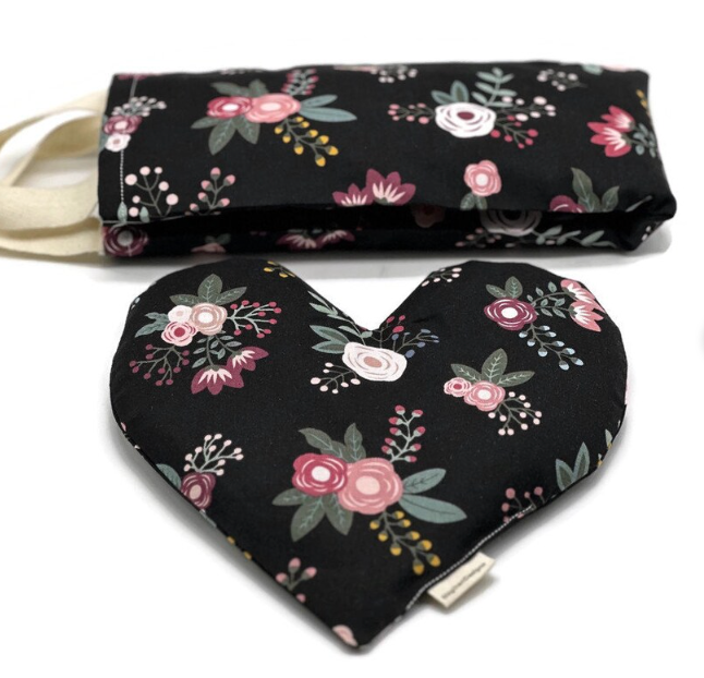 A matching set of handmade wellness products featuring a floral design on a black background. The set includes a rectangular eye pillow with a handle and a heart-shaped heating pad, both decorated with pink, white, and burgundy flowers