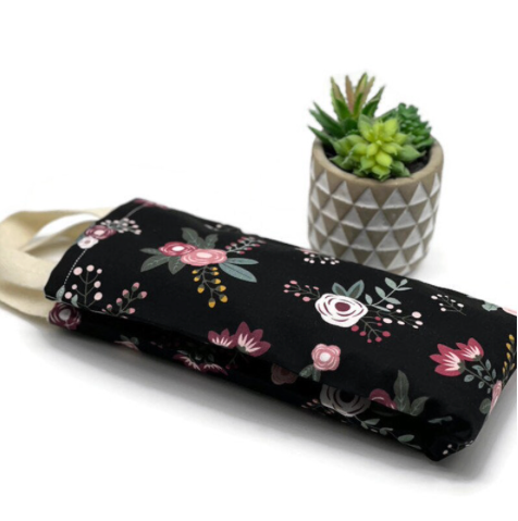 A handmade floral-patterned eye pillow with a handle, placed next to a small geometric pot containing a succulent plant. The eye pillow features a black background with pink, white, and burgundy flowers.