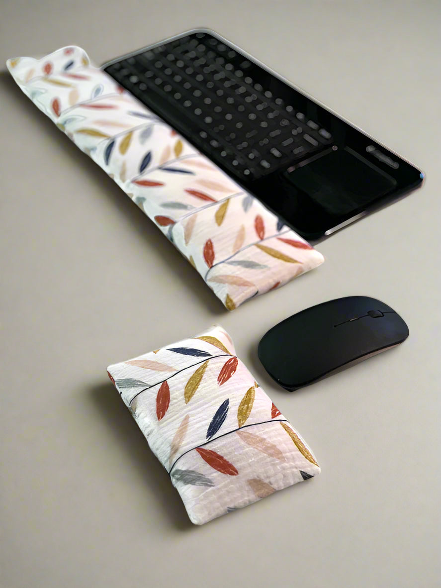 A black wireless keyboard with a built-in touchpad is placed on a white surface. In front of it lies a long, colorful leaves cream fabric wrist rest. Nearby, a white wireless mouse is accompanied by a smaller, matching wrist rest for ergonomic support.
