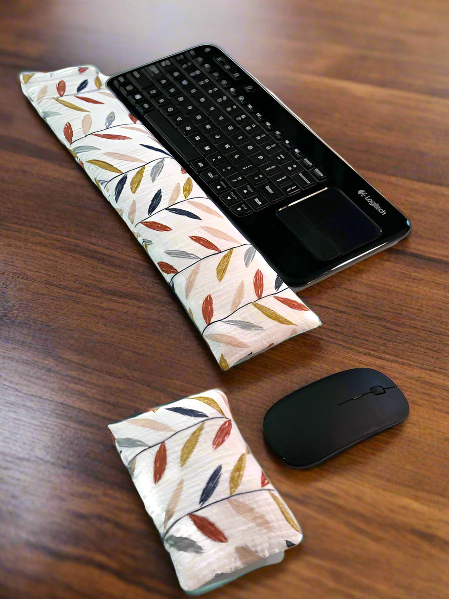 A black wireless keyboard with a built-in touchpad is placed on a white surface. In front of it lies a long, colorful leaves cream color fabric wrist rest. Nearby, a white wireless mouse is accompanied by a smaller, matching mouse wrist rest for ergonomic support.