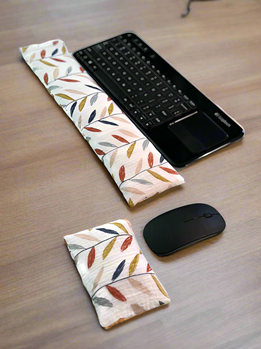 A black wireless keyboard with a built-in touchpad is placed on a white surface. In front of it lies a long, colorful leaves cream fabric wrist rest. Nearby, a white wireless mouse is accompanied by a smaller, matching fabric wrist rest for ergonomic support.