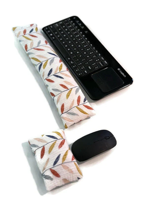 A black wireless keyboard with a built-in touchpad is placed on a white surface. In front of it lies a long, colorful leaves cream fabric wrist rest. Nearby, a white wireless mouse is accompanied by a smaller, matching fabric wrist rest for ergonomic support.
