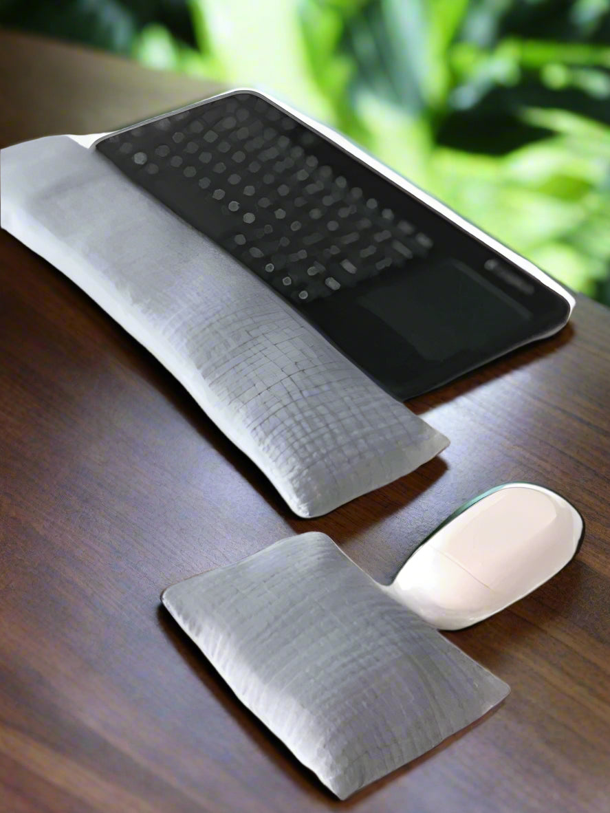 A black wireless keyboard with a built-in touchpad is placed on a white surface. In front of it lies a long, gray fabric wrist rest. Nearby, a white wireless mouse is accompanied by a smaller, matching gray wrist rest for ergonomic support.