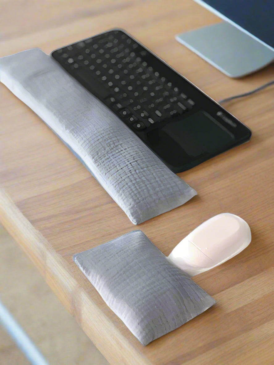 A black wireless keyboard with a built-in touchpad is placed on a white surface. In front of it lies a long, gray fabric wrist rest. Nearby, a white wireless mouse is accompanied by a smaller, matching gray wrist rest for ergonomic support.