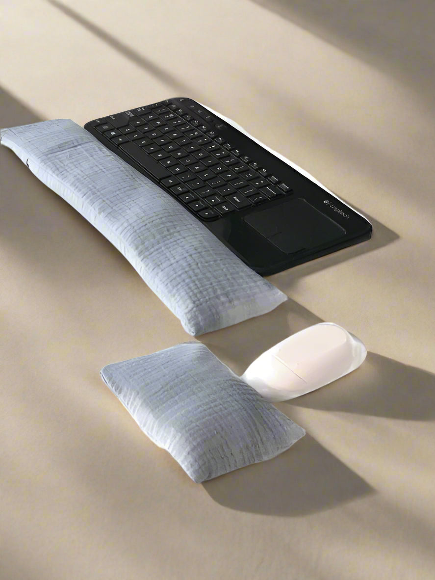 A black wireless keyboard with a built-in touchpad is placed on a white surface. In front of it lies a long, gray fabric wrist rest. Nearby, a white wireless mouse is accompanied by a smaller, matching gray wrist rest for ergonomic support.